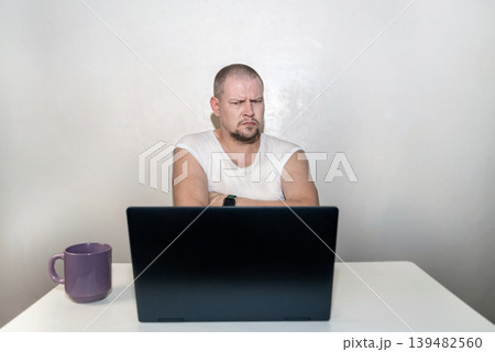 Thoughtful man with scowling face and crossed arms looking critically at a laptop screen, skeptical view Thoughtful man with scowling face and crossed arms looking critically at a laptop screen, skeptical view 139482560
