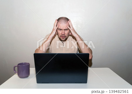 Stressed man clutches his head in shock and anger at an open laptop, experiencing frustration and despair Stressed man clutches his head in shock and anger at an open laptop, experiencing frustration and despair 139482561