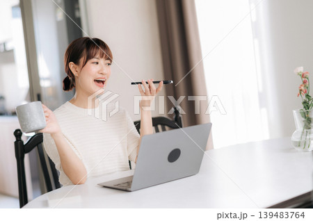 Happy Asian woman talking on smartphone using laptop at home desk holding coffee mug showing cheerful expression Happy Asian woman talking on smartphone using laptop at home desk holding coffee mug showing cheerful expression 139483764