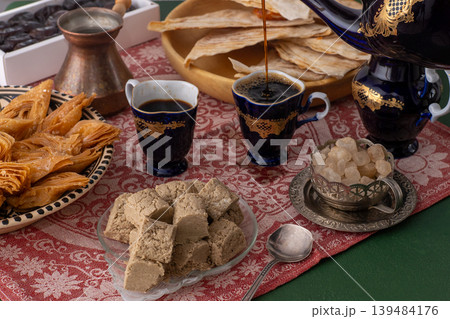 Pour coffee from a cobalt blue vintage decanter pot in cup. Sweets food on table honey baklava and halva on patterned red tablecloth 139484176