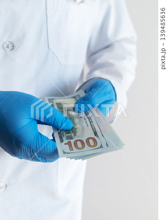 Close-up of doctor in white coat and blue gloves receiving or hiding cash. Concept of high medical salary, corruption in medicine, healthcare bribery, and expensive health insurance. Close-up of doctor in white coat and blue gloves receiving or hiding cash. Concept of high medical salary, corruption in medicine, healthcare bribery, and expensive health insurance. 139485636