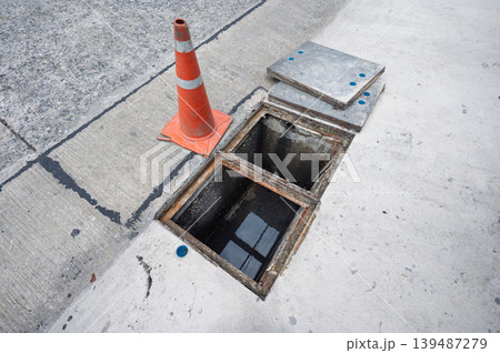 Scary open manhole hazard with orange traffic cone on concrete street 139487279