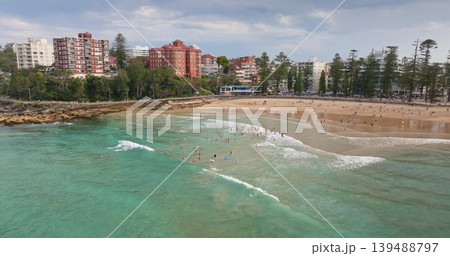 Panoramic aerial view people swimming in the turquoise Pacific Ocean water at Manly Beach, with sandy shores, bustling promenade, and coastal buildings lining the popular Sydney suburb. Drone panorama Panoramic aerial view people swimming in the turquoise Pacific Ocean water at Manly Beach, with sandy shores, bustling promenade, and coastal buildings lining the popular Sydney suburb. Drone panorama 139488797