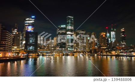 Sydney city skyline at night, modern skyscrapers with illuminated windows and vibrant urban landscape, reflecting colorful lights on tranquil harbor waters during the evening. Drone flight footage Sydney city skyline at night, modern skyscrapers with illuminated windows and vibrant urban landscape, reflecting colorful lights on tranquil harbor waters during the evening. Drone flight footage 139488806