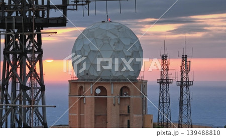 Large white radome and several telecommunication antennas standing on a hill overlooking the sea during a beautiful sunset, creating a striking silhouette against the colorful sky Large white radome and several telecommunication antennas standing on a hill overlooking the sea during a beautiful sunset, creating a striking silhouette against the colorful sky 139488810