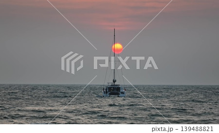 Catamaran vessel slowly sailing on the calm ocean water, traveling towards the glowing orange sun dropping below the horizon line during a beautiful golden hour sunset. Nature background 139488821