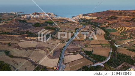 Malta, Comino island: Warm sunlight illuminating the landscape of Gozo, featuring cultivated fields, roads, and hotel buildings coastline in the distance. Aerial view drone flight Malta, Comino island: Warm sunlight illuminating the landscape of Gozo, featuring cultivated fields, roads, and hotel buildings coastline in the distance. Aerial view drone flight 139488849