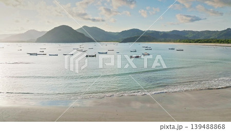 Selong Belanak Beach, Lombok: traditional fishing boats in calm bay with gentle waves and sandy shore, clear blue water and green hills under a sunny sky. Tropical nature landscape. Aerial view flight Selong Belanak Beach, Lombok: traditional fishing boats in calm bay with gentle waves and sandy shore, clear blue water and green hills under a sunny sky. Tropical nature landscape. Aerial view flight 139488868