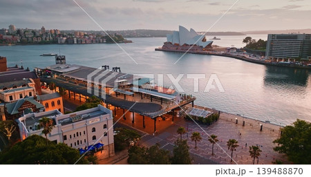 Sydney Opera House and Circular Quay waterfront at dramatic cloudy sunset dusk, ferries crossing the harbor and city skyline reflected in rippling water, iconic urban landmark. Aerial drone flight Sydney Opera House and Circular Quay waterfront at dramatic cloudy sunset dusk, ferries crossing the harbor and city skyline reflected in rippling water, iconic urban landmark. Aerial drone flight 139488870