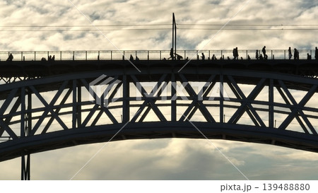 Silhouetted pedestrians people walking on the upper deck of Dom Luis I Bridge, spanning the Douro River in Porto, Portugal, at sunset, showcasing the iconic architecture and busy urban life 139488880