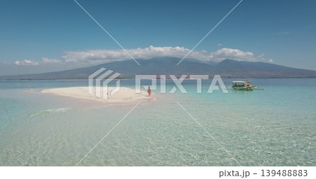 Woman walking on a pristine white sandbar surrounded by clear turquoise water, with a traditional longtail boat nearby and a volcanic mountain in the background of Lombok, Indonesia Woman walking on a pristine white sandbar surrounded by clear turquoise water, with a traditional longtail boat nearby and a volcanic mountain in the background of Lombok, Indonesia 139488883