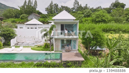 Woman drink coffe on modern luxury villa balcony enjoy beaytiful view on infinity swimming pool and lush green tropical landscape, relax, exclusive holiday vacation on Bali island. Aerial drone shot Woman drink coffe on modern luxury villa balcony enjoy beaytiful view on infinity swimming pool and lush green tropical landscape, relax, exclusive holiday vacation on Bali island. Aerial drone shot 139488886