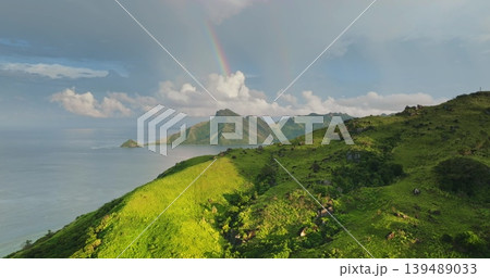 Fiji: Lush green mountains and hills meeting calm ocean waters with beautiful rainbow arching over the distant tropical islands and a cloudy sky. Perfect nature background. Aerial view drone flight Fiji: Lush green mountains and hills meeting calm ocean waters with beautiful rainbow arching over the distant tropical islands and a cloudy sky. Perfect nature background. Aerial view drone flight 139489033