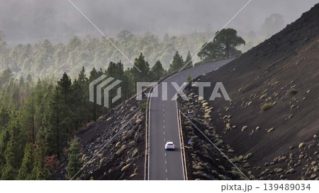 Tenerife, Canary Islands: Aerial view white car navigating winding road through volcanic landscape, with a misty forest backdrop near Mount Teide in Tenerife. Travel background. Drone flight Tenerife, Canary Islands: Aerial view white car navigating winding road through volcanic landscape, with a misty forest backdrop near Mount Teide in Tenerife. Travel background. Drone flight 139489034