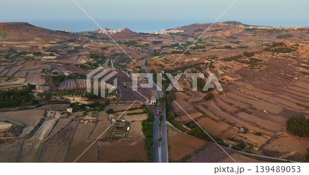 Malta, Comino island: Cultivated fields and a road crossing Gozo island with a red truck driving on it, aerial drone view during a sunny summer day in Malta. Hotel buildings coastline in background. Malta, Comino island: Cultivated fields and a road crossing Gozo island with a red truck driving on it, aerial drone view during a sunny summer day in Malta. Hotel buildings coastline in background. 139489053