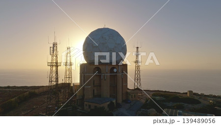 Large radar antenna and communication towers dominate the landscape of a coastal military installation, silhouetted against the warm hues of a setting sun over the calm sea. Drone flight 139489056