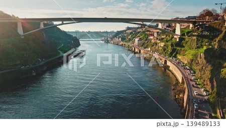 Portugal, Porto: Douro River with cruise boats, bridges and car traffic moving along the riverbank, showcasing urban city landscape with green hills under clear sky. Aerial view drone Portugal, Porto: Douro River with cruise boats, bridges and car traffic moving along the riverbank, showcasing urban city landscape with green hills under clear sky. Aerial view drone 139489133