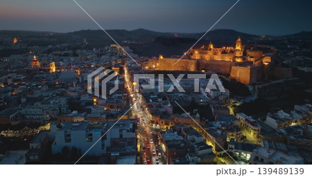 Malta, Comino island: Aerial view illuminated historic fortified Cittadella. Modern city buildings and streets glowing over Victoria on Gozo during enchanting twilight hours. Drone flight Malta, Comino island: Aerial view illuminated historic fortified Cittadella. Modern city buildings and streets glowing over Victoria on Gozo during enchanting twilight hours. Drone flight 139489139