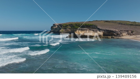 Penisok, Lombok, Indonesia: Ocean waves breaking on the sandy beach with rugged coastline cliff meeting the blue sky, creating a serene and natural tropical landscape. Wild nature. Aerial drone fligh 139489156