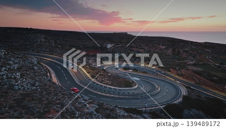 Malta: Winding road with cars driving along the coast of Malta during a beautiful sunset with pink and purple clouds, creating a scenic travel destination view. Aerial drone flight 139489172