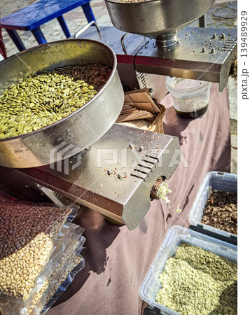 A stainless steel machine processes green pumpkin seeds on a table at an outdoor market stall. Finished oil drips from the machine while vendors prepare for customers. A stainless steel machine processes green pumpkin seeds on a table at an outdoor market stall. Finished oil drips from the machine while vendors prepare for customers. 139489929