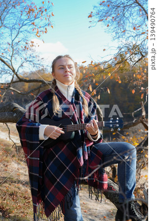 Blonde woman playing guitar ukulele outdoors in autumn forest. Concept of sound therapy, mental health and wellness rituals. Calmness tranquility audio-sensory practices. Aura farming 139490764