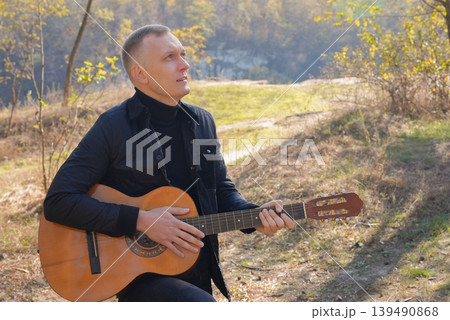 Beautiful blonde man playing acoustic guitar and singing relaxing enjoying outdoors on sunny autumn day. Artist learning and practicing playing musical instrument. Fall leaves autumnal Beautiful blonde man playing acoustic guitar and singing relaxing enjoying outdoors on sunny autumn day. Artist learning and practicing playing musical instrument. Fall leaves autumnal 139490868