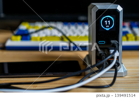A silver charging hub displaying 11W output, connected to a smartphone and other devices on a desk with a mechanical keyboard. A silver charging hub displaying 11W output, connected to a smartphone and other devices on a desk with a mechanical keyboard. 139491514