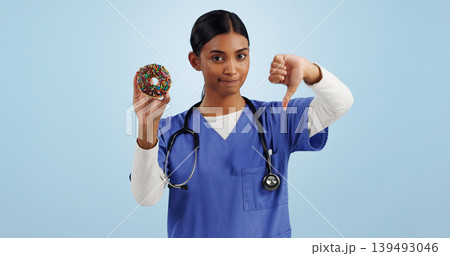 Woman, portrait and doctor with thumbs down, donut and bad review for diet on a blue studio background. Female person rejection, emoji or sign with chocolate dessert, diabetes and high blood sugar Woman, portrait and doctor with thumbs down, donut and bad review for diet on a blue studio background. Female person rejection, emoji or sign with chocolate dessert, diabetes and high blood sugar 139493046