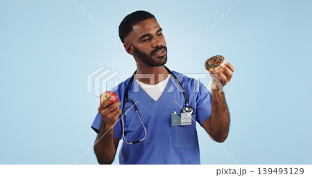 Doctor, man and studio for donut, apple and choice with thinking for healthcare by blue background. Medic, person or nutritionist with decision, cake and fruit with ideas for food, eating and diet Doctor, man and studio for donut, apple and choice with thinking for healthcare by blue background. Medic, person or nutritionist with decision, cake and fruit with ideas for food, eating and diet 139493129