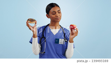 Apple, donut and nurse in studio with choice for healthy eating, diet and detox on blue background. Thinking, healthcare and Indian woman with dessert, fruit and cake for decision, option and balance Apple, donut and nurse in studio with choice for healthy eating, diet and detox on blue background. Thinking, healthcare and Indian woman with dessert, fruit and cake for decision, option and balance 139493169