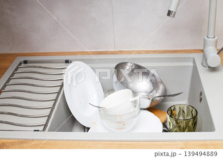 Pile of dirty dishes like plates, cutlery in the grey modern granite sink in the kitchen Pile of dirty dishes like plates, cutlery in the grey modern granite sink in the kitchen 139494889