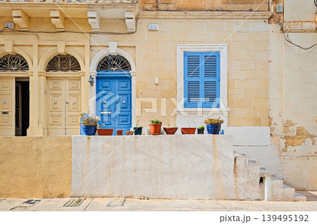Sunny facade of historic Malta limestone building with bright blue wooden door, matching shutters, ornate ironwork arch, terracotta and blue flower pots on white wall 139495192