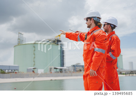 Engineers in orange uniforms inspecting LNG storage tanks at coastal terminal under cloudy sky Engineers in orange uniforms inspecting LNG storage tanks at coastal terminal under cloudy sky 139495685