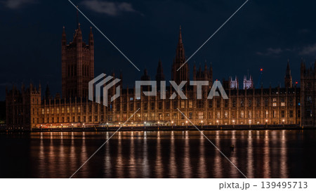 Illuminated gothic building stands along river with reflections under dark sky. Repeating windows and lights create a detailed historic architecture scene. 139495713