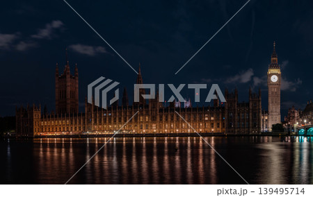 Illuminated clock tower and long gothic building stand beside river under dark sky. Warm lights reflect on water forming clear architectural night scene. Illuminated clock tower and long gothic building stand beside river under dark sky. Warm lights reflect on water forming clear architectural night scene. 139495714