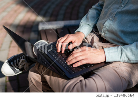 Man sitting on bench and working on laptop. Freelancer typing on keyboard in park. Digital nomad lifestyle, remote work and business communication in urban city environment concept. Man sitting on bench and working on laptop. Freelancer typing on keyboard in park. Digital nomad lifestyle, remote work and business communication in urban city environment concept. 139496564