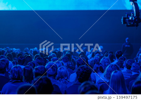 Audience seated in blue lit conference hall watching presentation, crowded rows at live event 139497175