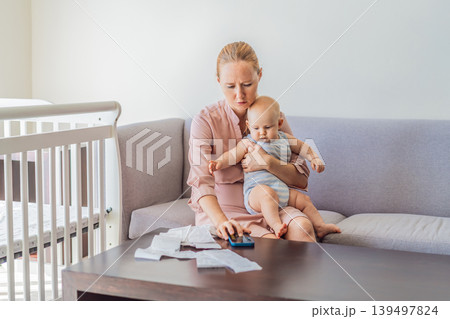Mother sitting on sofa holding baby while calculating monthly expenses with receipts at home. Stressed mom managing budget and childcare. Financial stress, parenthood and real life concept Mother sitting on sofa holding baby while calculating monthly expenses with receipts at home. Stressed mom managing budget and childcare. Financial stress, parenthood and real life concept 139497824