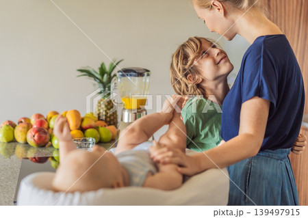 Mother and son preparing smoothie in kitchen while baby lies in cozy lounger on table watching. Family cooking together, healthy lifestyle, parenting, sibling bonding and everyday life concept Mother and son preparing smoothie in kitchen while baby lies in cozy lounger on table watching. Family cooking together, healthy lifestyle, parenting, sibling bonding and everyday life concept 139497915