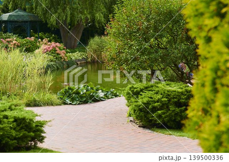 Serene garden path winding beside tranquil pond with lush greenery and colorful blooms Serene garden path winding beside tranquil pond with lush greenery and colorful blooms 139500336