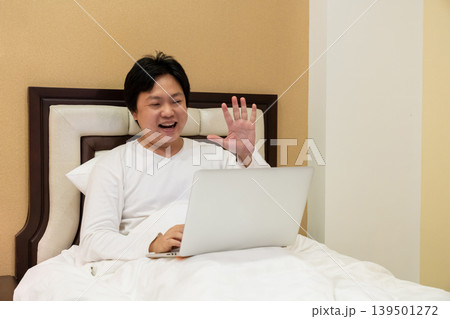 The portrait of a young man leans back against the bed waving hand to a person, friend, family in the video call. Using a laptop to contact with others on a bed. The portrait of a young man leans back against the bed waving hand to a person, friend, family in the video call. Using a laptop to contact with others on a bed. 139501272