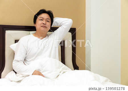 A tired Asian man placed his palm behind his neck, suffering from shoulder and neck pain, sitting up in a bed leaning back on a pillow in a bedroom. 139501278
