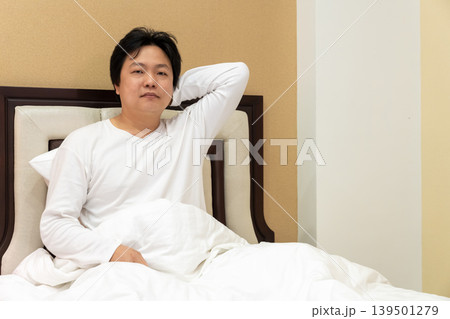 A tired Asian man placed his palm behind his neck, suffering from shoulder and neck pain, sitting up in a bed leaning back on a pillow in a bedroom. 139501279