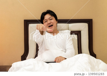 Asian man on bed pointing toward camera with playful expression, combining casual lifestyle gesture with direct viewer engagement and personal connection. Asian man on bed pointing toward camera with playful expression, combining casual lifestyle gesture with direct viewer engagement and personal connection. 139501283