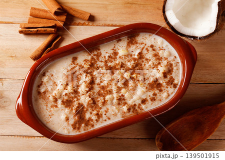 A bowl of creamy white corn porridge with cinnamon, accompanied by peanut brittle and coconut, served on a wooden base. A popular traditional Brazilian A bowl of creamy white corn porridge with cinnamon, accompanied by peanut brittle and coconut, served on a wooden base. A popular traditional Brazilian 139501915