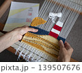 Close-up of a person weaving a handmade tapestry with lighthouse motif on a wooden frame loom. Weaver following a paper sketch to create a textile landscape with colorful wool yarns 139507676