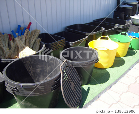 Household and gardening equipment. It features a variety of durable black plastic tubs, vibrant yellow and green flexible buckets, and a collection of traditional straw brooms with colorful handles 139512900