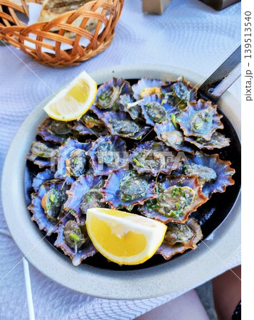 A dish featuring several grilled limpets, Patella spp. served in a round ceramic bowl, garnished with two lemon wedges. A woven basket with slices of bread A dish featuring several grilled limpets, Patella spp. served in a round ceramic bowl, garnished with two lemon wedges. A woven basket with slices of bread 139513540