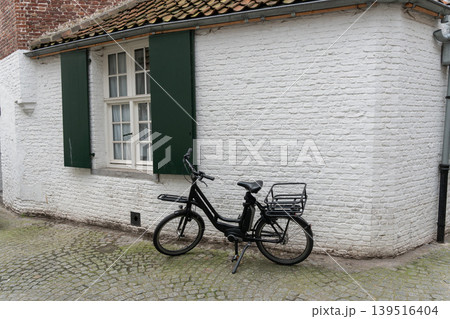 Black bicycle stands beside white brick house with green shutters, tiled roof, cobblestone pavement, quiet street mood in Bruges. Cycling culture, city travel, sustainable mobility, architecture Black bicycle stands beside white brick house with green shutters, tiled roof, cobblestone pavement, quiet street mood in Bruges. Cycling culture, city travel, sustainable mobility, architecture 139516404